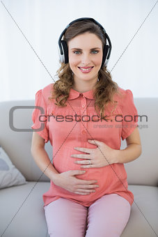 Cute pregnant woman sitting on couch while relaxing and listening to music