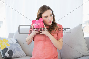 Lovely pregnant woman shaking a piggy bank sitting on couch
