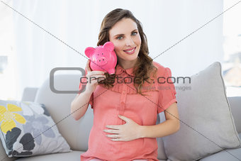 Content pregnant woman shaking pink piggy bank sitting on couch