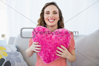 Cheerful pregnant woman holding pink heart pillow sitting on couch