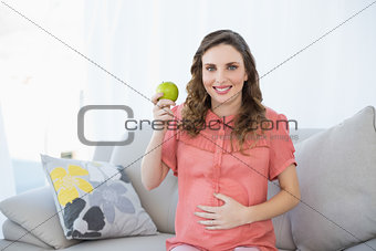 Young pregnant woman showing green apple sitting on couch in living room
