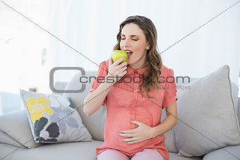 Lovely pregnant woman eating green apple sitting on couch in living room