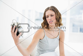 Beautiful annoyed woman holding an alarm clock looking at it sitting on her bed
