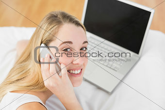 Beautiful woman phoning while lying on her bed next to her notebook