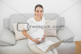 Smiling pregnant woman sitting on couch using her laptop touching her belly