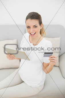Beautiful pregnant woman holding her credit card and tablet looking cheerfully at camera