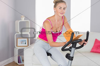 Sporty smiling blonde training on exercise bike reading a book