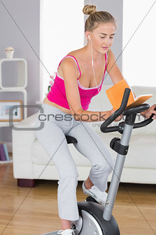 Sporty happy blonde training on exercise bike reading a book