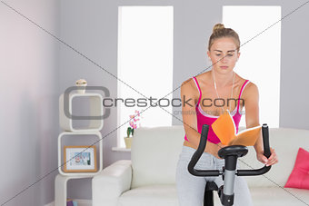 Sporty focused blonde training on exercise bike reading a book