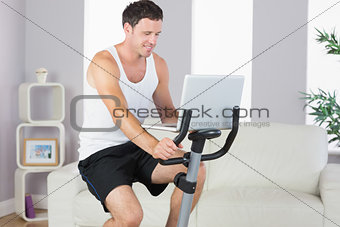 Content sporty man exercising on bike and using laptop