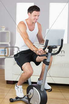 Happy sporty man exercising on bike and using laptop