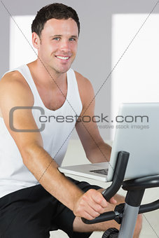 Attractive sporty man exercising on bike and holding laptop