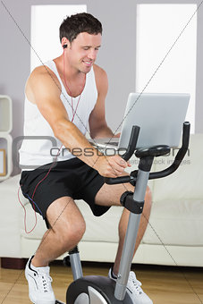 Sporty man with earphones exercising on bike looking at laptop