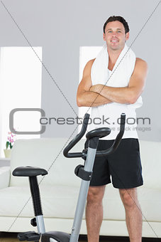 Smiling sporty man standing next to exercise bike cross armed