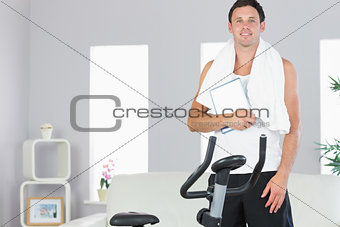 Smiling sporty man standing behind exercise bike holding tablet