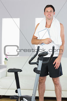 Cheerful sporty man standing behind exercise bike holding tablet