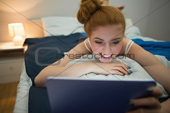 Happy redhead using digital tablet lying on her bed