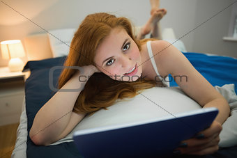 Smiling redhead using digital tablet lying on her bed