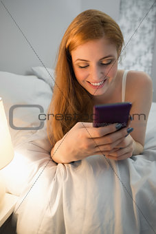 Cheerful redhead lying on bed sending a text