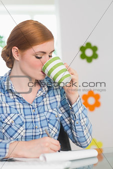 Redhead writing on notepad at her desk and drinking coffee