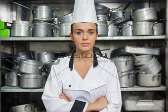 Young content chef standing arms crossed between shelves