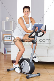 Happy training woman using an exercise bike while holding a tablet
