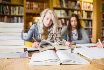 Students writing notes with stack of books at library desk
