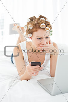 Shocked woman in hair curlers with cellphone looks at laptop in bed