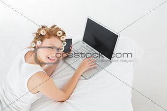 Cheerful woman in hair curlers using laptop in bed
