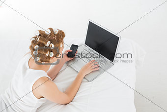 Woman in hair curlers with cellphone using laptop in bed
