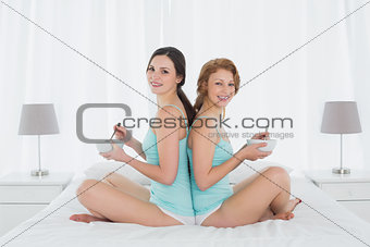 Smiling female friends with salad bowls sitting back to back on bed