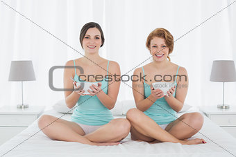 Smiling female friends with salad bowls sitting on bed
