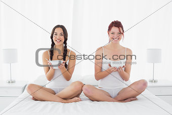 Smiling female friends with bowls sitting on bed