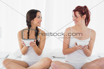 Smiling young female friends with bowls sitting on bed