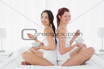 Two smiling female friends with bowls sitting on bed