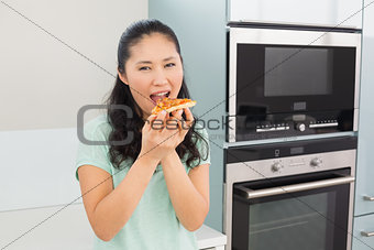 Smiling young woman eating a slice of pizza in kitchen