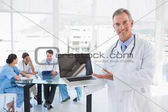 Doctor holding laptop with group around table in hospital