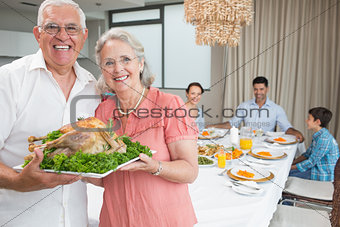 Grandparents holding chicken roast with family at dining table