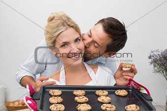 Man kissing woman's cheek as she holds freshly baked cookies