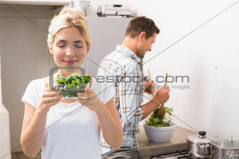 Woman holding bowl of leaves with man preparing salad in background in kitchen