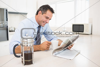 Smiling well dressed man with coffee cup reading newspaper in kitchen