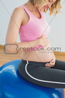 Happy blonde pregnant woman touching belly on exercise ball