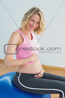 Smiling blonde pregnant woman touching belly on exercise ball