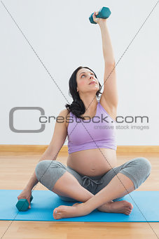 Pregnant brunette sitting on exercise mat lifting hand weight