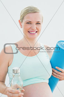 Pregnant woman holding bottle of water and exercise mat smiling at camera