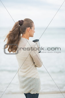 Young woman standing on cold beach with cup of hot beverage. rea