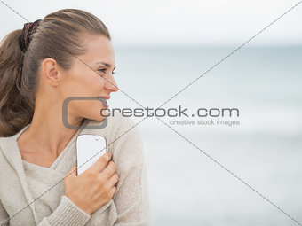 Young woman with cell phone standing on cold beach and looking o