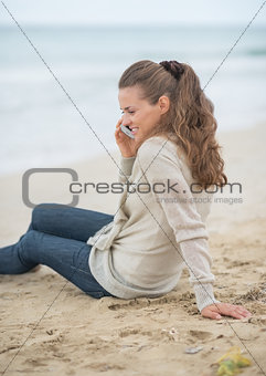 Young woman sitting on cold beach and talking mobile phone