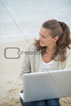 Young woman sitting with laptop on cold beach and looking into d