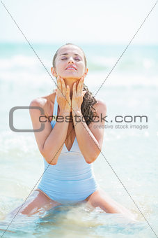 Young woman in swimsuit sitting on sea shore and washing face